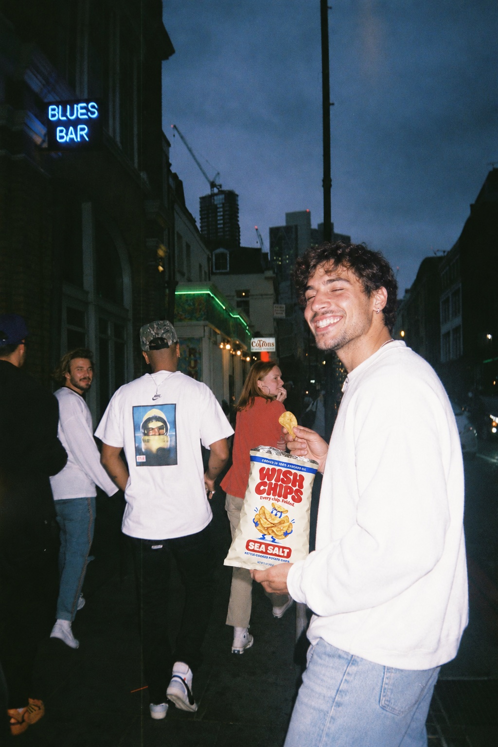 A smiling man holds a chip and a Wish Chips bag outside a blues bar with friends.
